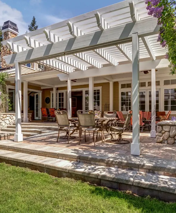 A beautiful, spacious stone patio featuring a large, white attached pergola, built by professional pergola builders to extend the home's outdoor living space and provide filtered shade.