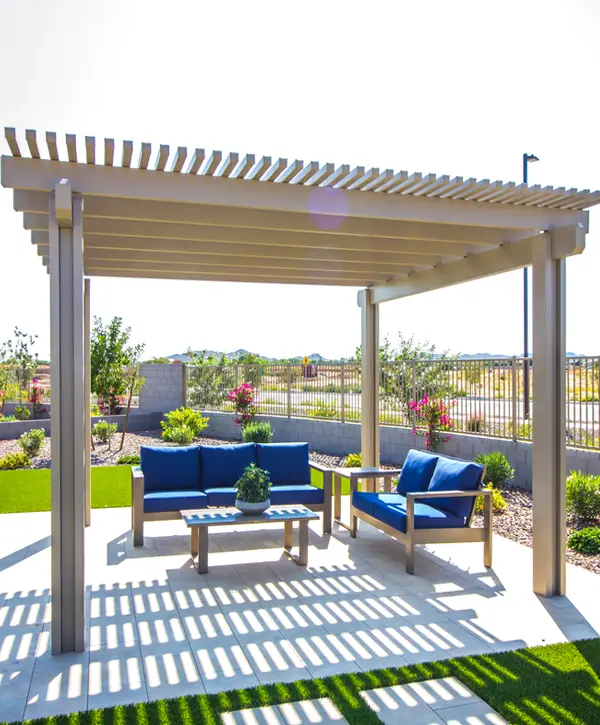 A modern tan louvered pergola standing over a paved patio area featuring a blue three-seater sofa, a matching loveseat, and a wooden coffee table in a sunlit backyard garden.
