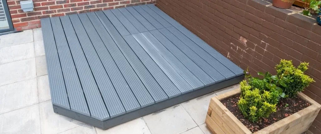 A modern grey composite ground level deck installed over a stone tile patio next to a brick wall and a wooden planter box with green shrubs.