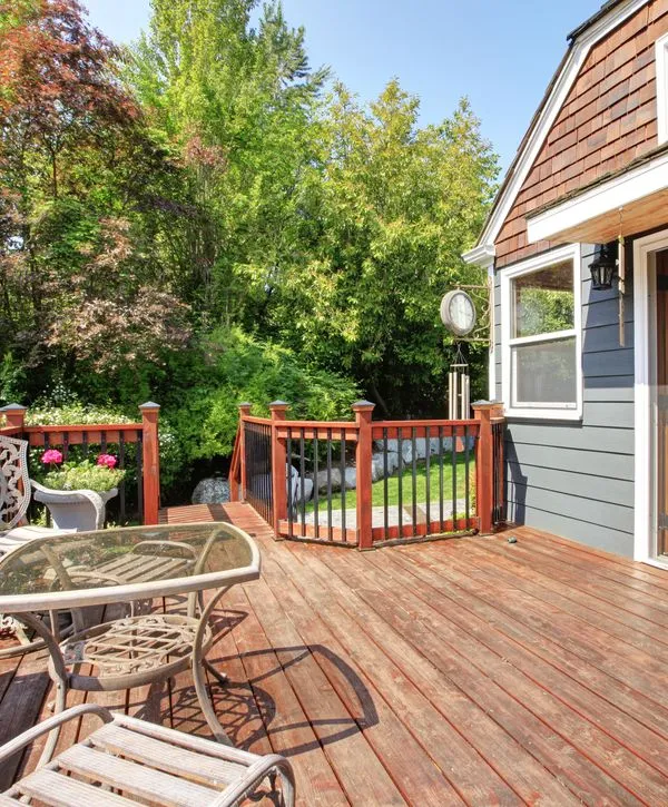 A sunlit wooden backyard deck built by professional deck builders in Cicero, IN, featuring a patio table, chairs, and a safety railing overlooking lush greenery.