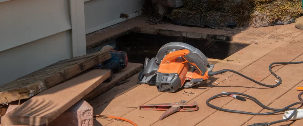 An orange circular saw sitting on a wooden deck next to a cut opening intended for an under deck drainage installation.