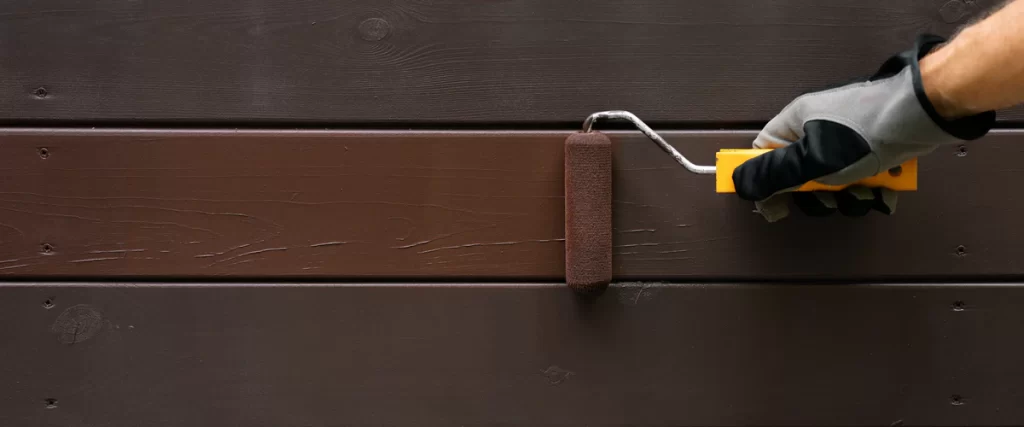 A person wearing a protective glove using a small paint roller to apply smooth brown paint to horizontal wood planks.