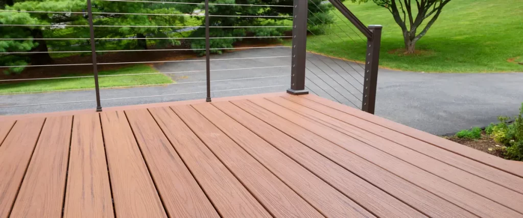 A finished brown wood-grain composite deck featuring a modern dark metal cable railing system overlooking a lush green backyard.