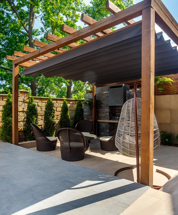 A modern dark wood pergola installed by Lawrence, IN pergola builders featuring a retractable black fabric shade over a stone patio with comfortable wicker seating and a hanging egg chair.