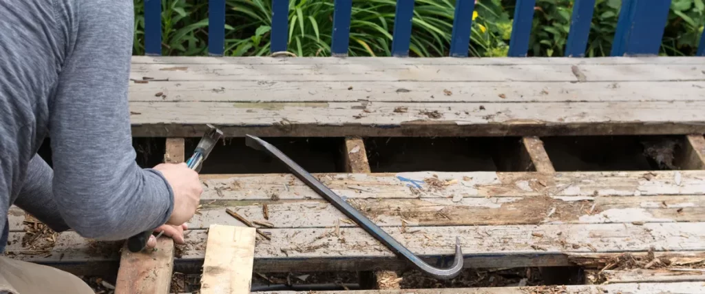 A person using a hammer and a crowbar to pry up old, peeling painted wooden deck boards, exposing the structural joists beneath.