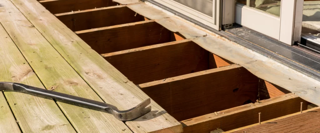A black crowbar resting on old green treated deck boards next to a removed section that reveals the wooden deck joist framing near a glass door.