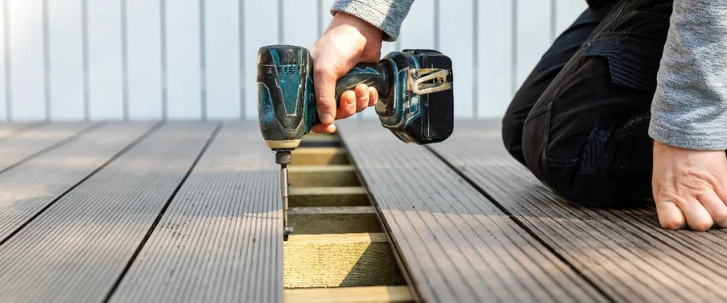 A person kneeling on a deck surface and using a blue cordless power drill to fasten grey grooved composite decking boards onto a wooden joist substructure.